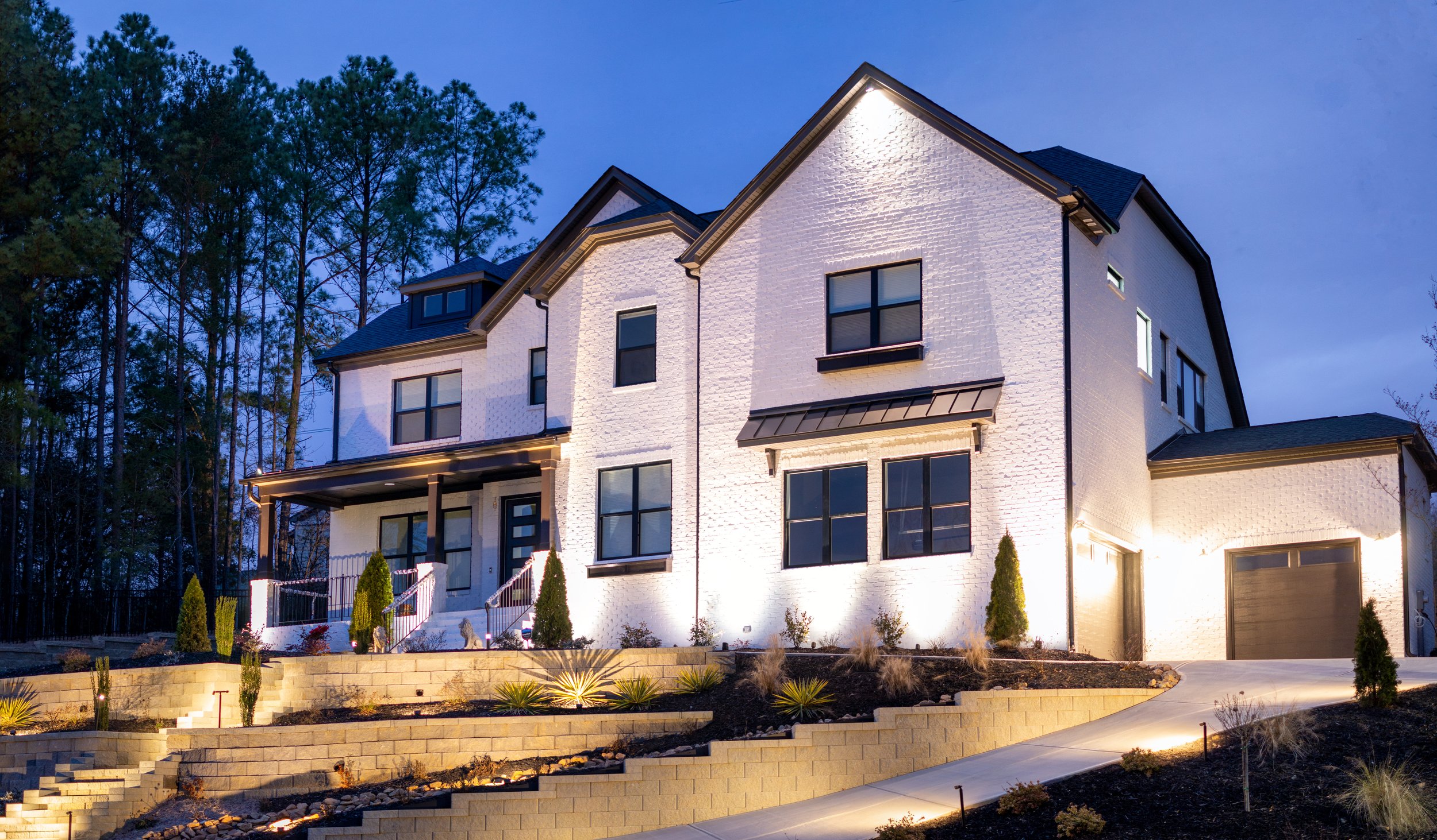 White brick home with dark trim illuminated with architectural up lights and downlights. Front yard is heavily landscaped with tiered retaining walls illuminated with soft landscape lighting. 