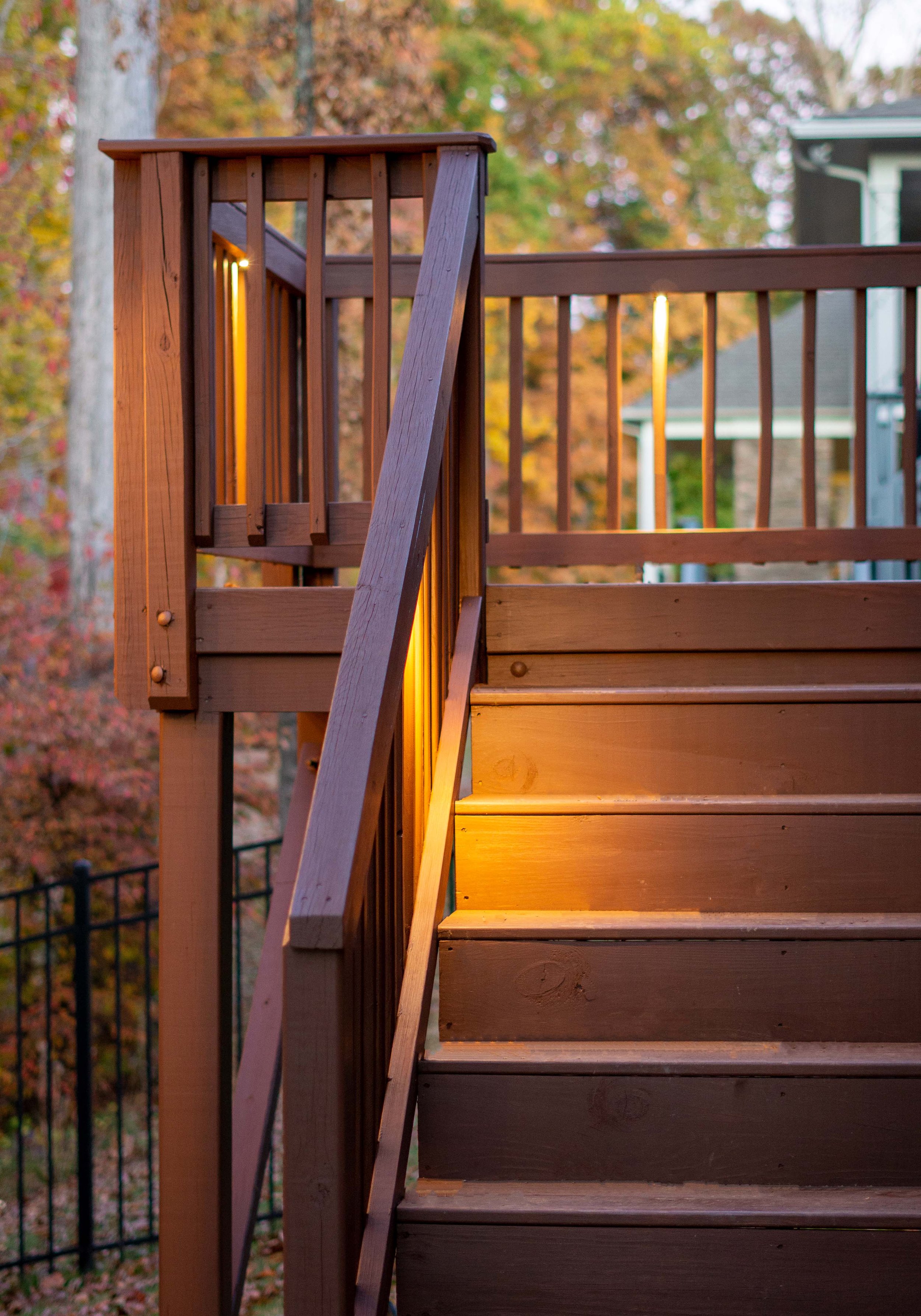 Image featuring deck lighting. Brown wooden stairs leads up to a deck with lights mounted under the railings. Fall trees can be seen in the background.
